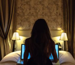 Woman sitting on a bed facing a laptop in a softly lit bedroom, preparing for a video call.