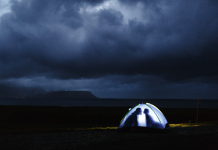 A small illuminated tent on a dark shore, with two silhouetted figures inside facing each other, beneath heavy storm clouds over distant mountains and sea.