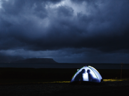 A small illuminated tent on a dark shore, with two silhouetted figures inside facing each other, beneath heavy storm clouds over distant mountains and sea.