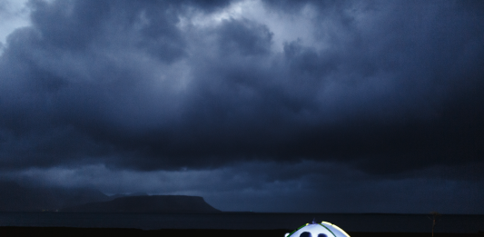 A small illuminated tent on a dark shore, with two silhouetted figures inside facing each other, beneath heavy storm clouds over distant mountains and sea.