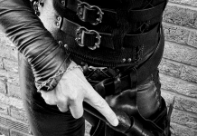 FemDom Mistress Cara in laced leather corset and trousers, holding a punishment tool at Her side, photographed from waist down against a brick wall