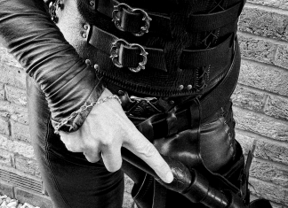 FemDom Mistress Cara in laced leather corset and trousers, holding a punishment tool at Her side, photographed from waist down against a brick wall