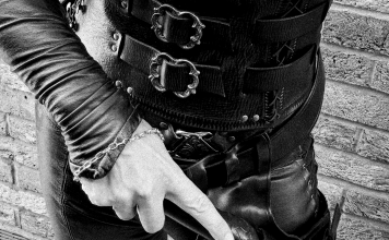 FemDom Mistress Cara in laced leather corset and trousers, holding a punishment tool at Her side, photographed from waist down against a brick wall