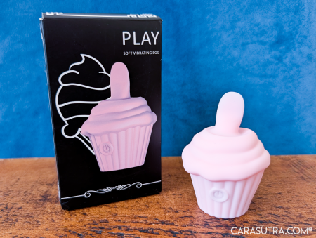 Pink Cupcake Vibrator on a wooden surface beside its black PLAY box against a blue backdrop