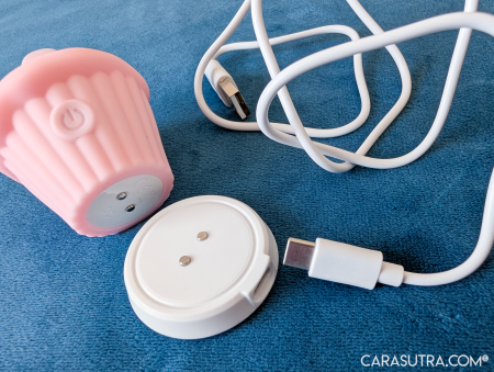 Pink Cupcake Vibrator lying on its side next to its white circular magnetic charging dock and USB-C wire on blue velvet, showing the metal contact points on the base of the toy