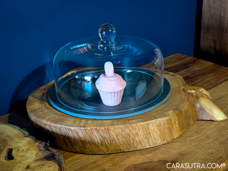 Pink Cupcake Vibrator displayed under a clear glass cake dome on a round wooden platter, styled like a patisserie display piece against a deep blue wall