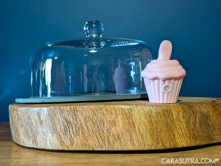 Pink Cupcake Vibrator positioned beside a clear glass cake dome on a round wooden platter, showing how discreetly it blends in with kitchen homeware