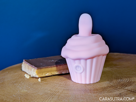 Pink Cupcake Vibrator standing upright on a wooden surface beside a slice of caramel shortbread, illustrating the toy's cupcake styling with a real cake treat