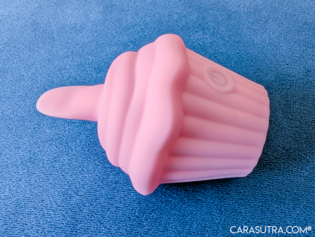 Pink Cupcake Vibrator lying on its side against blue carpet, showing the opposite side view with the power button visible