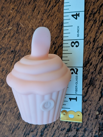 Pink Cupcake Vibrator on a dark wood surface with a white measuring tape held vertically alongside it for height reference