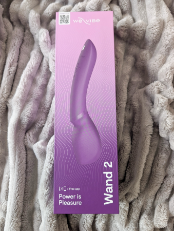 We-Vibe Wand 2 packaging front on a grey blanket, showing the Power is Pleasure branding and product silhouette through the box design