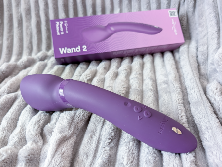 We-Vibe Wand 2 vibrator on a grey blanket next to the mostly purple Wand 2 packaging
