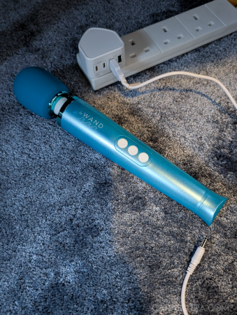 Le Wand Dive vibrator on a dark blue carpet next to its plugged in charging wire, plugged into a USB to mains plug