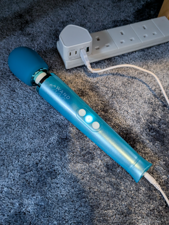 Le Wand Dive vibrator on a dark blue carpet with its charging wire plugged into it and the central operation button lit up white, plugged into a USB to mains plug