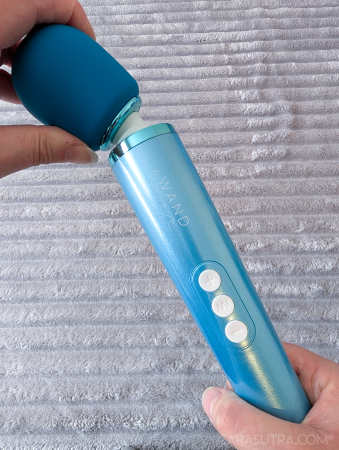 Hand holding the Le Wand Dive vibrator upright, showing the flex of the silicone head at the neck, the chrome collar and three white control buttons on the tapered handle