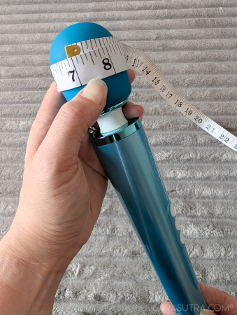 Cara holds the Le Wand Dive vibrator in her hands with a grey fluffy blanket, in the background. A measuring tape is around the widest part of the head, showing maximum circumference as 7 inches