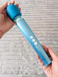 Cara holds the Le Wand Dive vibrator in her two hands, showing its full self in a vertical pose, portrait oriented photo. in the background is a light grey fluffy blanket. 