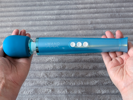 Le Wand Dive submersible wand vibrator held horizontally in two hands against a light grey fluffy blanket, showing the full length, silicone head, three white control buttons and tapered handle for scale