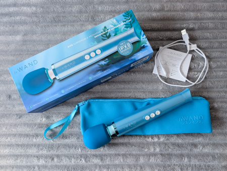 flat lay of unboxed Le Wand Dive vibrator with box contents, on a light grey fluffy blanket. Box, the wand vibrator, storage bag, charging wire and instruction booklet laid out. 