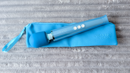 Le Wand Dive wand vibrator lying next to its matching blue zip-up storage and travel bag with wrist strap on a grey fluffy blanket
