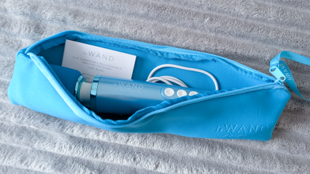 Le Wand Dive submersible wand vibrator inside its open blue zip-up storage bag alongside the coiled charging cable and instruction booklet, on a grey ribbed background