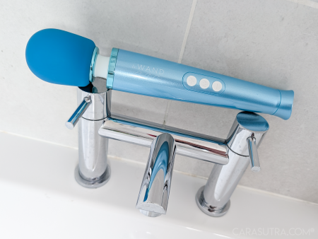 Le Wand Dive wand vibrator resting on top of chrome bath taps against a white tiled bathroom wall