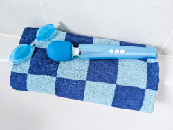 Le Wand vibrator resting on a light blue and dark blue checked towel which is folded on the side of a white bath. Beside the wand vibe is a pair of light blue swimming goggles. 