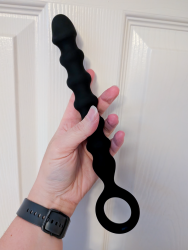 Cara's hand holds a sizeable anal probe against a white door. It's a black, beaded Titus Anal Probe with a finger loop at the base.