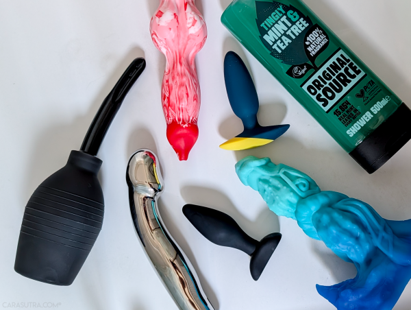 Flat lay of anal douching kit and toys on white background: black silicone bulb anal douche, chrome metal G-spot dildo, red and white marbled fantasy dildo, small dark blue silicone butt plug with yellow base, black classic silicone butt plug, turquoise and blue fantasy dildo, and Original Source Tingly Mint & Tea Tree shower gel bottle