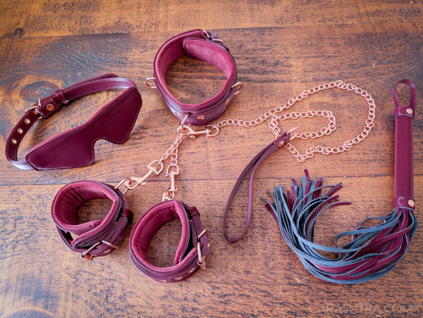 Liebe Seele Metallic Shades Vegan Bondage Set in burgundy, showing collar with leash, wrist cuffs, blindfold and flogger arranged on a wooden surface