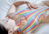 Cara Sutra photographed from above, lying on a cream fleece throw wearing a rainbow striped cutout lingerie bodysuit, one hand resting on her stomach