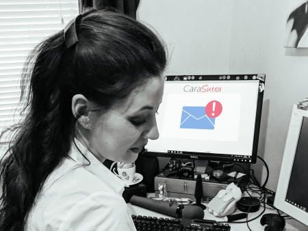 Cara Sutra at her office desk looking at a computer screen showing the CaraSutra logo and an email alert icon, with vibrators beside the keyboard