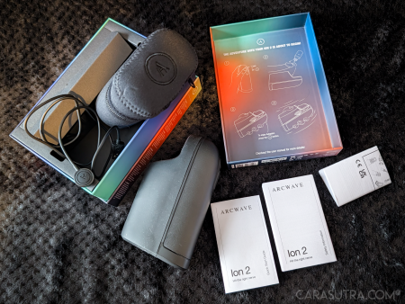 Arcwave Ion 2 unboxing flatlay on a black blanket, with the open box tray showing the charging cable and zip case, the stroker, the Quick Start Guide and Safety Information booklets, and the inner lid printed with the “The adventure with your Ion 2 is about to begin!” setup diagram