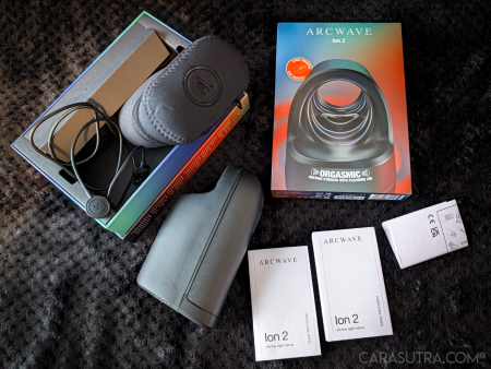 Arcwave Ion 2 full unboxing flatlay on a black blanket, showing the open retail box with the magnetic charging cable coiled inside, the black neoprene zip case, the black stroker, the upright product box, and the Quick Start Guide and Safety Information booklets plus the folded UK/CE leaflet