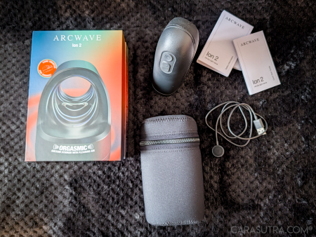 Arcwave Ion 2 unboxing flatlay with the retail box, stroker, zip case, charging cable and quick start guides arranged on a black blanket